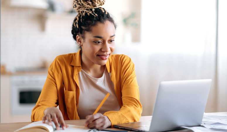 A woman making notes beside a laptop at a kitchen table