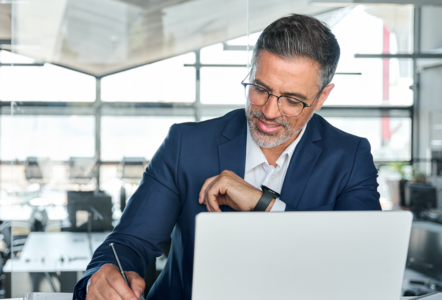 Mid aged business man working on laptop computer in office writing notes.