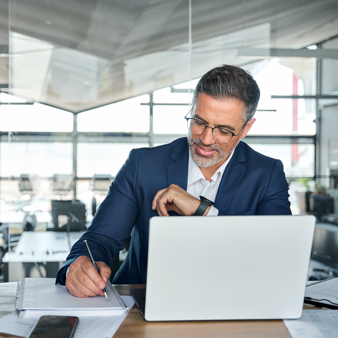 Mid aged business man working on laptop computer in office writing notes.