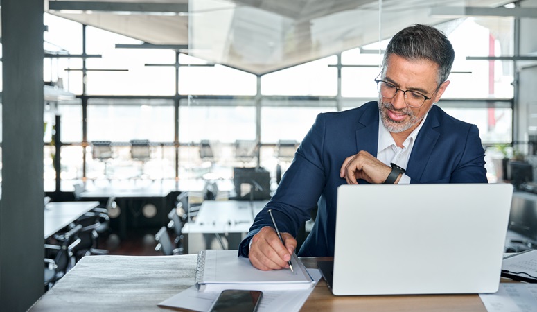 Mid aged business man working on laptop computer in office writing notes.